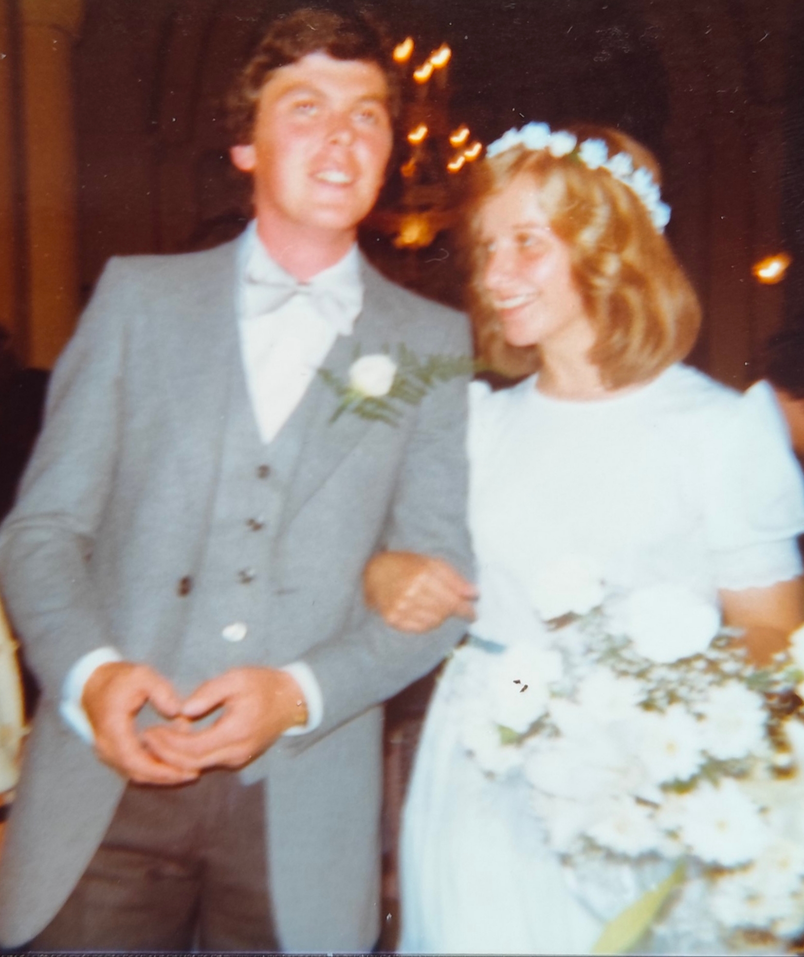 Les mariés le jour de leur mariage, elle en robe blanche avec une couronne de fleurs, lui en costume gris.